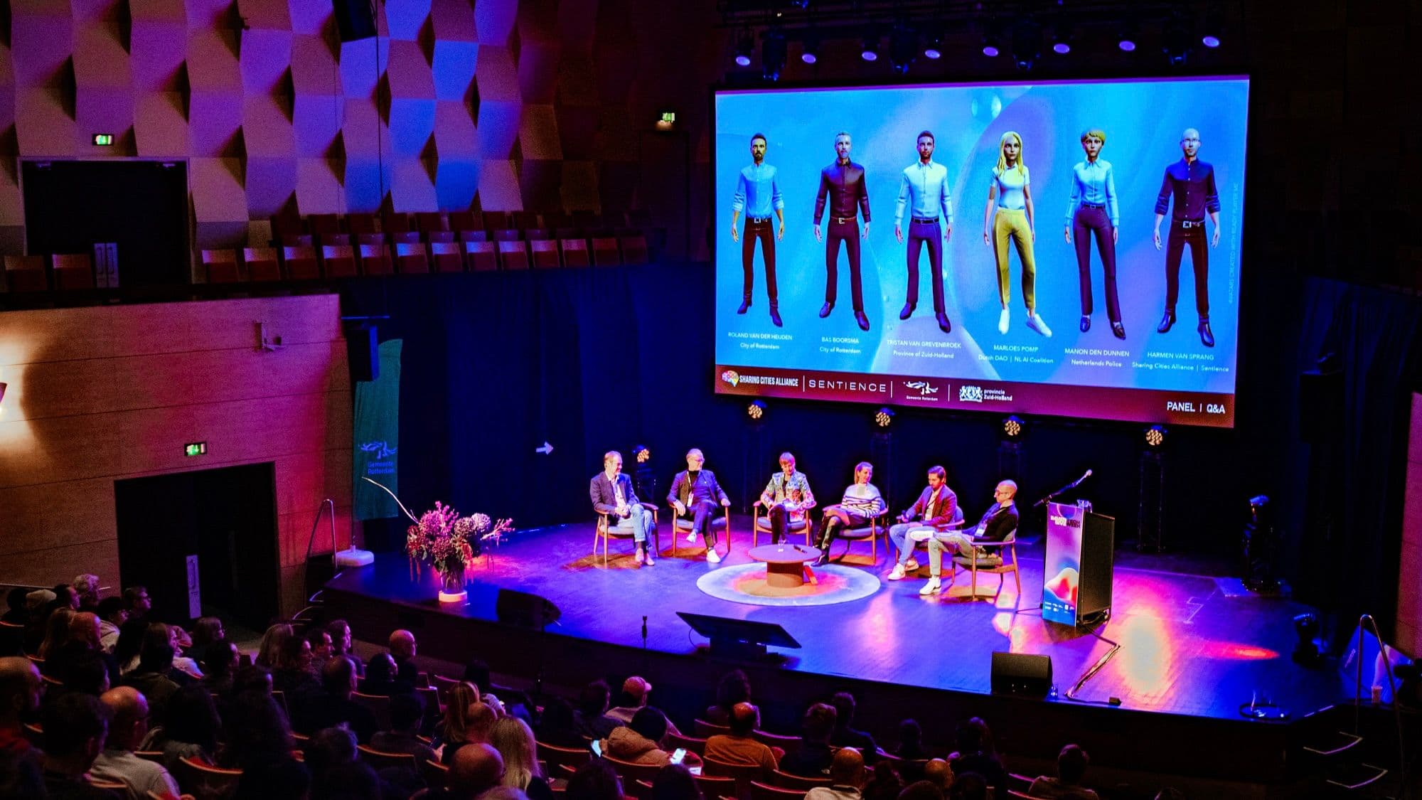 A group of six people having a discussion on a stage at Immersive Tech Week, above them a big screen feat. six avatars | by JaynoBrk