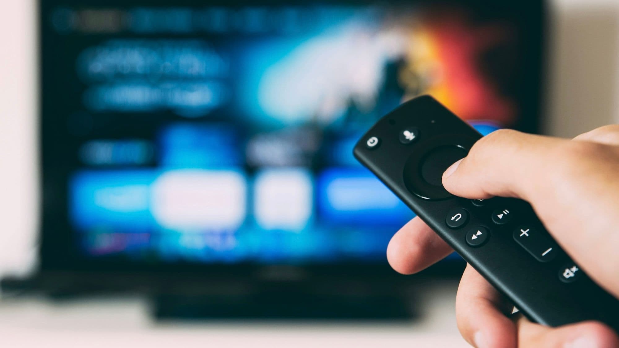 Photo of a person holding a smart TV remote, with a TV screen in the background (by Glenn Carstens-Peters, Unsplash)