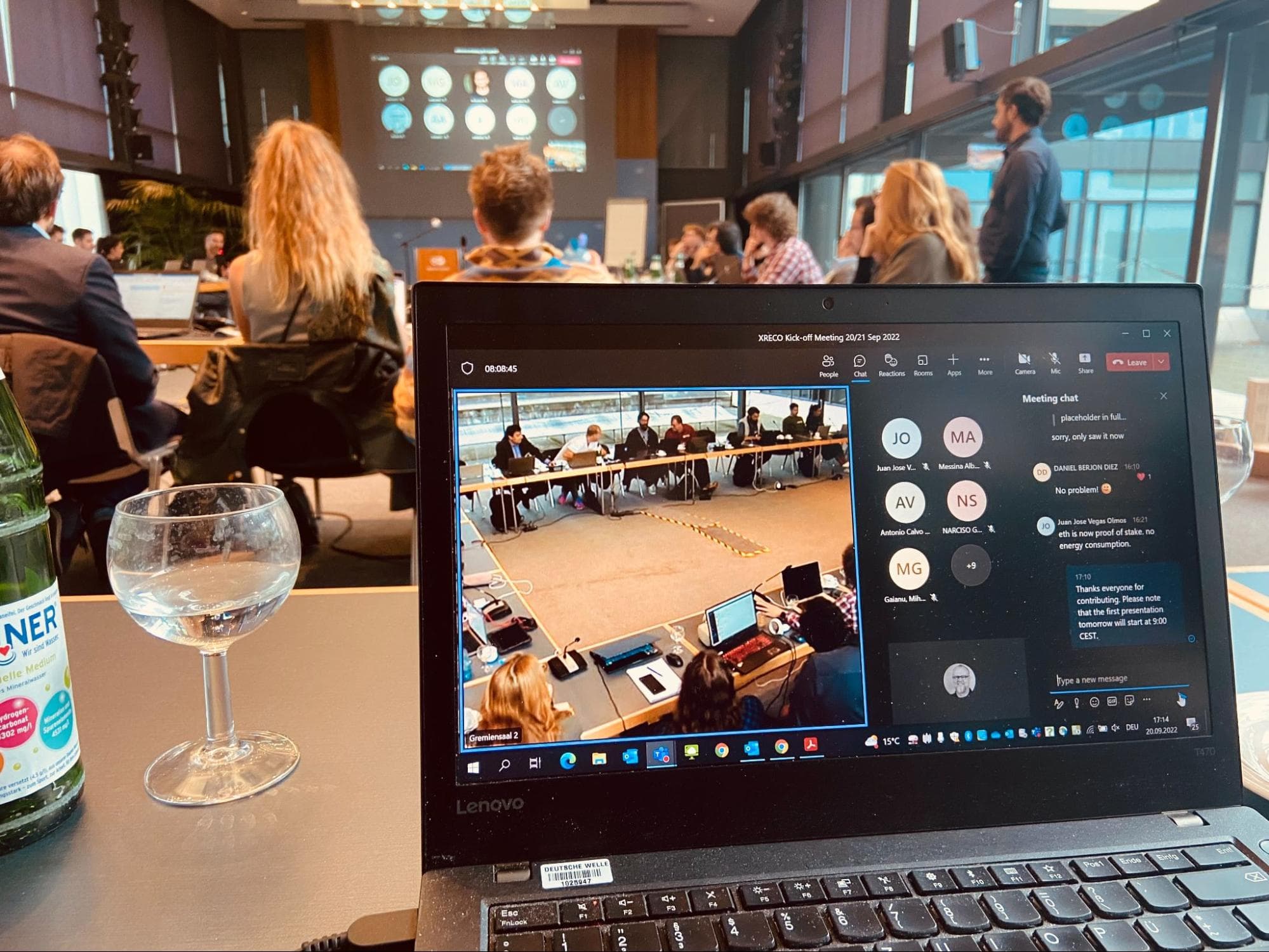 Picture shows a large group of diverse people gathered at a table in a conference room in the background – and the same scene from a different angle on a laptop in the foreground.