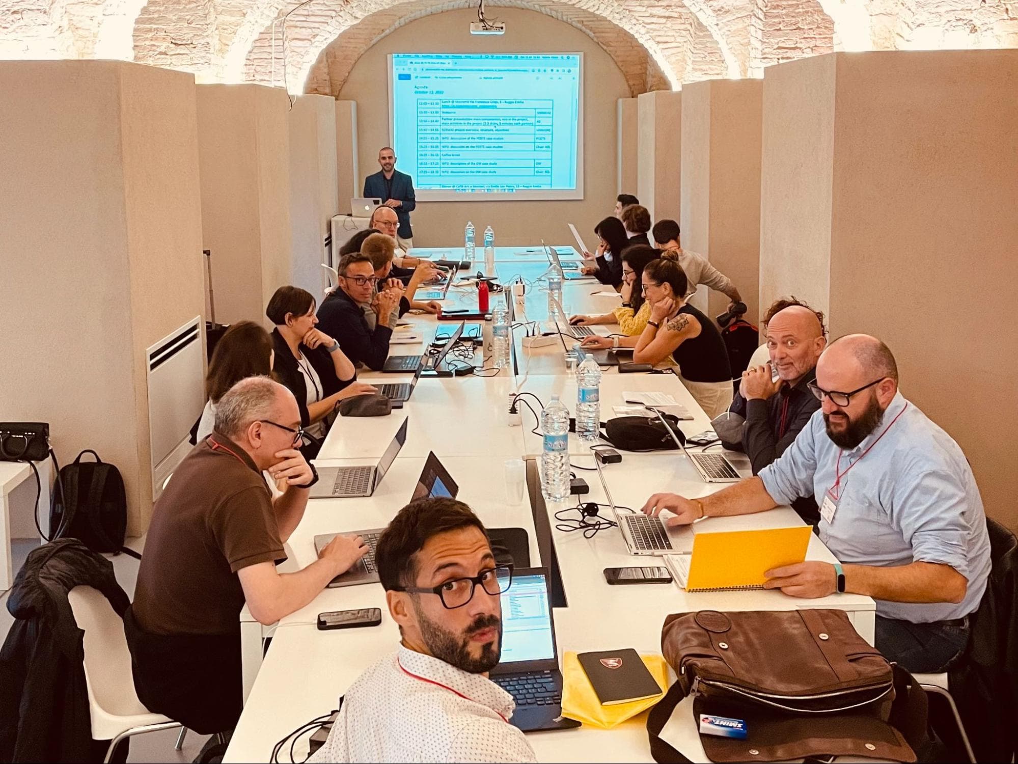 Picture shows a large group of diverse people sitting and working at a table under an old stone vault; some of them are looking into the camera.