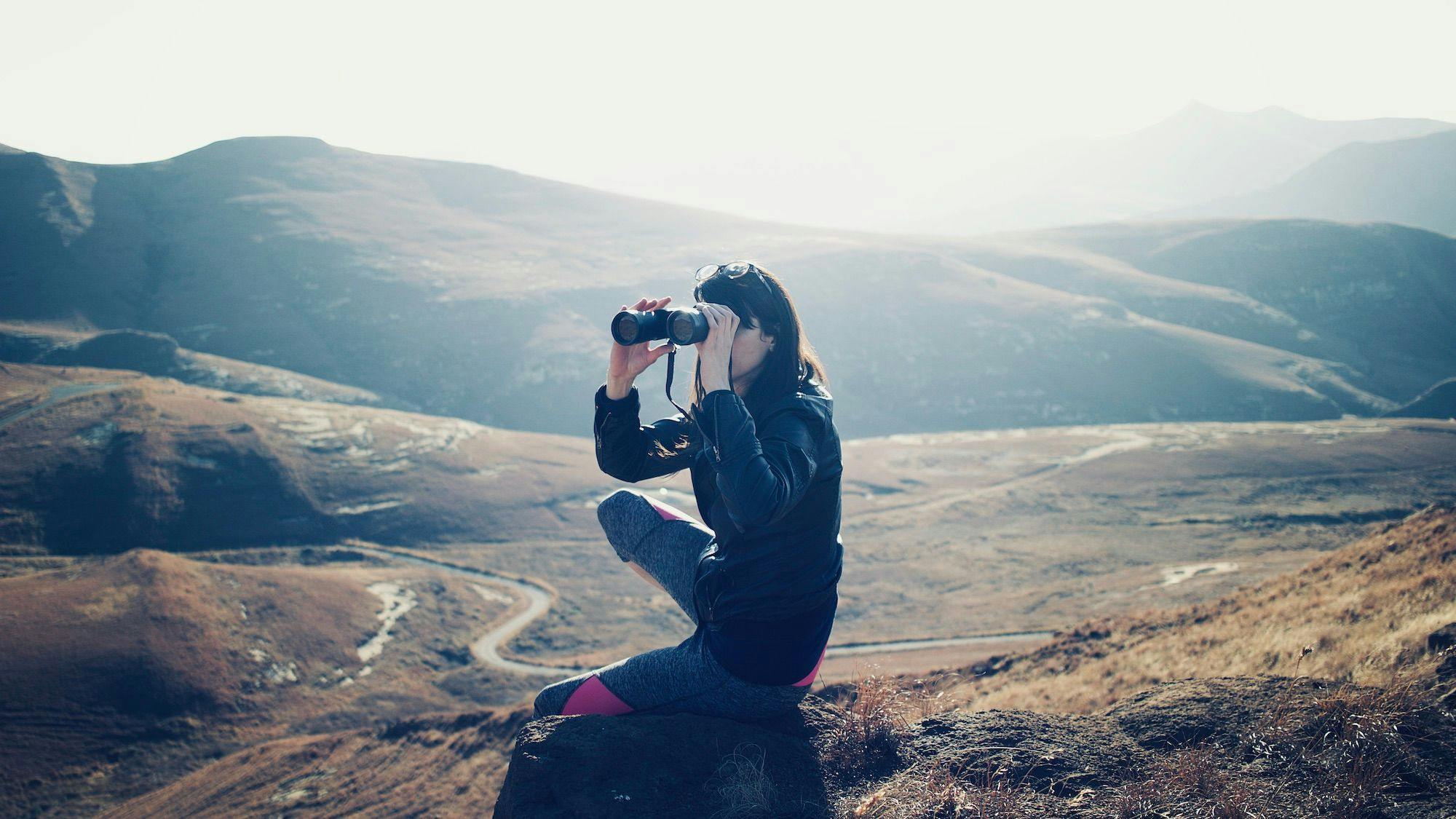 Photo of a woman sitting on a rock and looking through binoculars