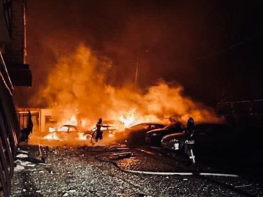 Picture shows the results of a Russian bombardment on the outskirts of Kharkiv. It's dark. Several cars and a building are on fire. There's debris everywhere. Two people seem to be fighting the fire.